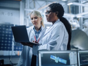 Two Female Engineers Talk, Use Computer working at Aerospace Satellite Manufacturing Facility. Diverse Team of Multi-Ethnic Scientists Doing Science Technology Research for Space Exploration Program